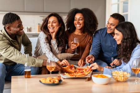 Foto de Un grupo diverso de amigos multirraciales adultos jóvenes riendo junto con bebidas y comida durante una cómoda fiesta en casa - Imagen libre de derechos