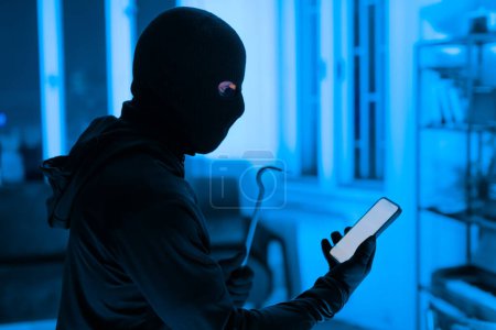 The image depicts a focused thief utilizing a smartphone to aid in a burglary, emphasizing the modern tools used in crime