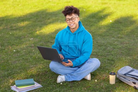 Photo for Smiling brazilian guy student casually studying with a laptop on the grass, surrounded by books and a backpack in a park-like campus - Royalty Free Image