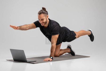 Téléchargez les photos : Un homme athlétique faisant un exercice de planche devant un ordinateur portable sur un tapis de yoga, signifiant le mélange de la santé et de la technologie dans la forme physique personnelle - en image libre de droit