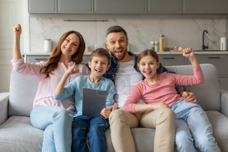 Foto de Una familia sonriente con dos niños sentados juntos en un sofá, levantando alegremente los puños con una tableta digital en las manos - Imagen libre de derechos