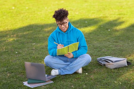 Téléchargez les photos : Un étudiant brésilien englouti dans les notes écrites dans un carnet tout en étant assis sur l'herbe avec un ordinateur portable à côté de lui sur le campus - en image libre de droit