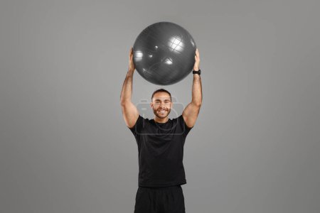 Foto de Un hombre sonriente levanta una gran pelota de fitness por encima de su cabeza mostrando fuerza y salud sobre fondo gris - Imagen libre de derechos