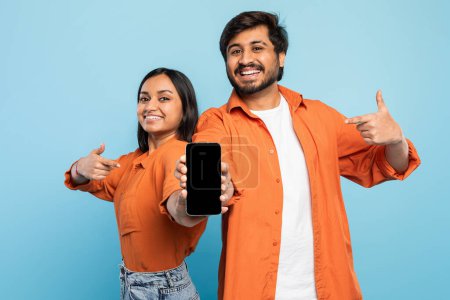 Indisches Paar lächelt selbstbewusst und zeigt mit dem Daumen auf ein Smartphone, perfekt für Technologie-Anzeigen