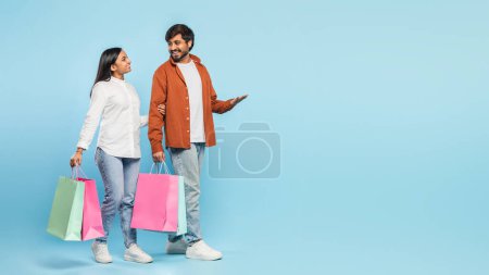Liebendes hinduistisches Paar, das Einkaufstüten trägt, tauscht Blicke aus und genießt einen gemeinsamen Moment vor blauem Hintergrund