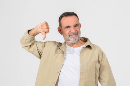 Photo for Disgruntled older man with a beard giving a thumbs down sign, signifying disapproval or dissatisfaction on white backdrop - Royalty Free Image