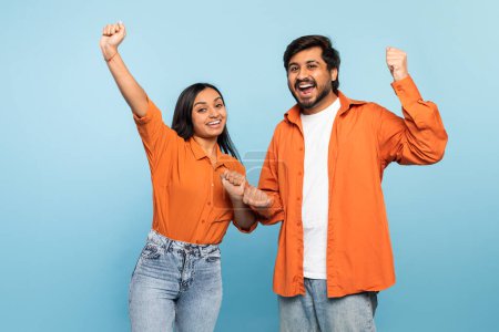 Energetisches indisches Paar mit erhobenen Fäusten feiert einen Sieg oder Erfolg in passender orangefarbener Kleidung vor blauem Hintergrund