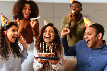 Foto de Un alegre grupo de amigos multirraciales canta la canción de cumpleaños a un amigo sosteniendo un pastel con velas encendidas, celebrando juntos - Imagen libre de derechos
