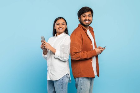 Foto de Hombre y mujer indios se paran espalda con espalda sonriendo a la cámara mientras ambos sostienen los teléfonos inteligentes sobre un fondo azul - Imagen libre de derechos