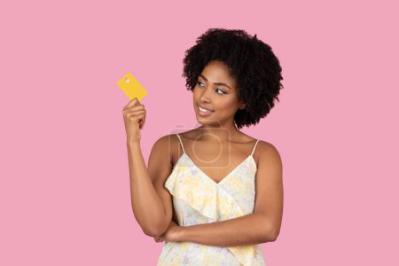 Thoughtful African American woman holding a yellow credit card, looking away on pink studio background