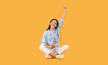 Cheerful young lady celebrating a success with a raised fist while sitting against an orange backdrop