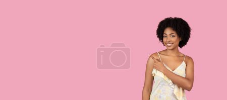 Foto de Una joven y alegre mujer afroamericana con el pelo rizado apuntando hacia un lado sobre un fondo rosa, espacio para copiar - Imagen libre de derechos