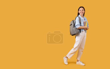 Confident young student walking with a backpack and a stack of books on a bright yellow background, copy space