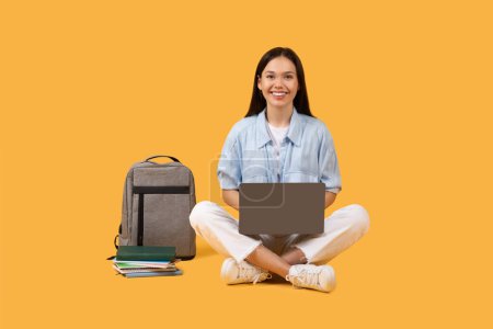 Joyeux jeune étudiante avec un ordinateur portable et un sac d'école assis par terre sur un fond jaune
