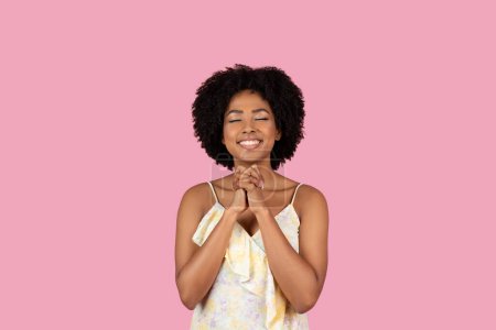Téléchargez les photos : Joyeux jeune afro-américaine avec les cheveux bouclés tenant ses mains ensemble, souriant avec les yeux fermés sur un fond rose - en image libre de droit