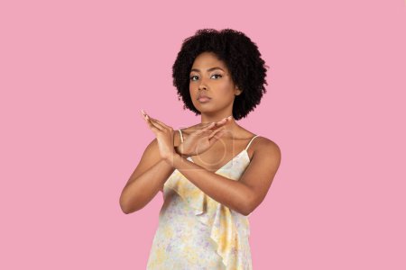 A young African American woman with curly hair in a floral dress stands against a pink background, crossing her hands in an X shape to signal stop or refusal