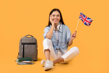 Junge Frau mit Kopfhörern, die eine britische Flagge in der Hand hält, sitzt neben einem Rucksack und Büchern