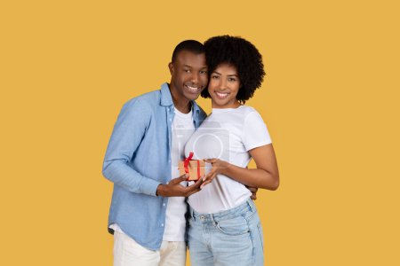 Zärtliches afroamerikanisches Paar hält ein verpacktes Geschenk in der Hand und lächelt, symbolisiert das gemeinsame Glück