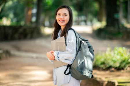 Junge glückliche Studentin hält Laptop in der Hand und steht im sonnigen Außenbereich eines öffentlichen Parks