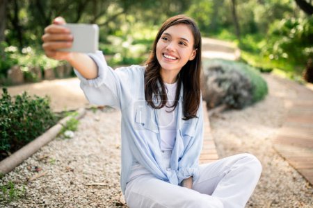Foto de Mujer joven emocionada captura una selfie en un parque, lo que refleja la tendencia de la autodocumentación y las redes sociales - Imagen libre de derechos