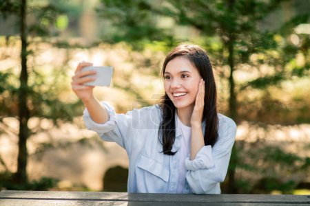 Femme heureuse prend un selfie avec son téléphone dans la nature, souriant et touchant ses cheveux au parc public