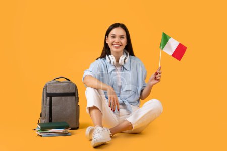 Joven alegre mostrando una bandera italiana, acompañada de auriculares y material educativo