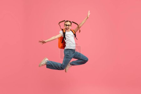 Photo for A cheerful girl wearing a backpack jumps with arms raised high against a pink background, showing happiness - Royalty Free Image