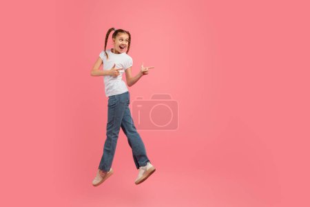 Photo for Energetic young girl in jeans and a white shirt jumps and points to the side on a pink background, copy space - Royalty Free Image