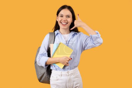 Téléchargez les photos : Une jeune femme joyeuse avec un sac à dos, tenant un carnet de notes fait semblant de faire un geste d'appel téléphonique sur un fond jaune - en image libre de droit