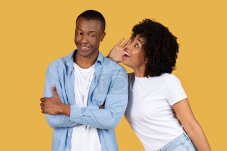 African American young woman is whispering into a mans ear, suggesting they are sharing a secret on a yellow background