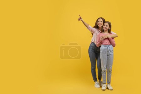 Téléchargez les photos : Duo souriant, mère et jeune fille pointant quelque chose ci-dessus avec excitation sur fond jaune, espace de copie - en image libre de droit