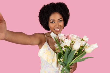 Foto de Sonriente joven afroamericana tomando una selfie con un hermoso ramo de flores, telón de fondo rosa - Imagen libre de derechos