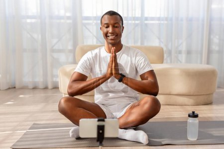 A relaxed African American man in sportswear sits cross-legged practicing yoga, recording with smartphone on a tripod