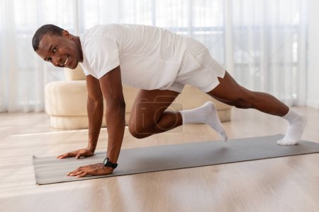 African american man in a white outfit holds a strong plank position on a yoga mat within a bright, spacious room