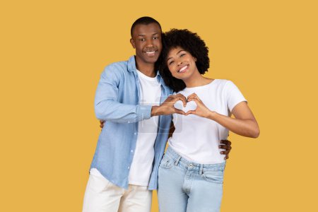 Téléchargez les photos : Jeune couple afro-américain souriant et faisant une forme de coeur avec leurs mains, symbolisant l'amour sur fond jaune - en image libre de droit