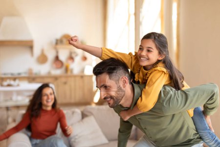 Photo for A playful family scene with a father giving his daughter a piggyback ride as the mother watches on happily - Royalty Free Image