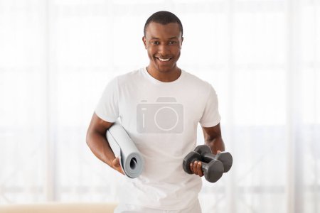 Hombre negro sonriente en camiseta blanca equipado con una esterilla de yoga y pesas para una sesión de entrenamiento