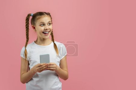 Photo for Excited young girl holding a smartphone looks away on a pink background showing curiosity and anticipation - Royalty Free Image
