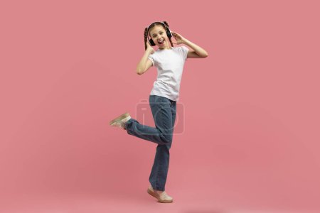 Photo for Young teen girl with headphones dances with one leg up against a pink backdrop showing fun and freedom - Royalty Free Image