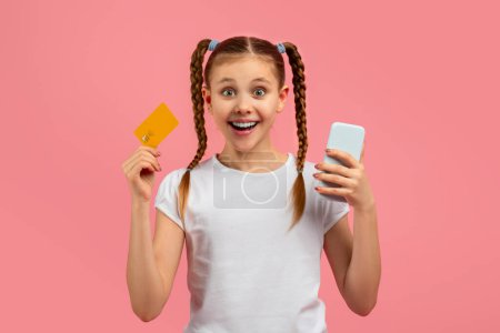Foto de Una joven encantada sosteniendo una tarjeta de crédito y un teléfono inteligente con una expresión alegre en el fondo rosa - Imagen libre de derechos