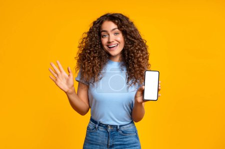 Foto de Mujer sonriente que presenta una pantalla de teléfono inteligente en blanco mientras saluda felizmente con su otra mano, aislada en amarillo - Imagen libre de derechos