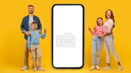 Foto de Familia alegremente posando de cuatro al lado de una pantalla de teléfono inteligente en blanco para maqueta de espacio publicitario sobre fondo amarillo - Imagen libre de derechos