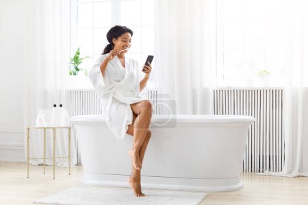 Foto de Una elegante mujer afroamericana con una túnica blanca disfruta de su mañana sentada en el borde de una bañera, interactuando con su teléfono inteligente, cepillándose los dientes - Imagen libre de derechos
