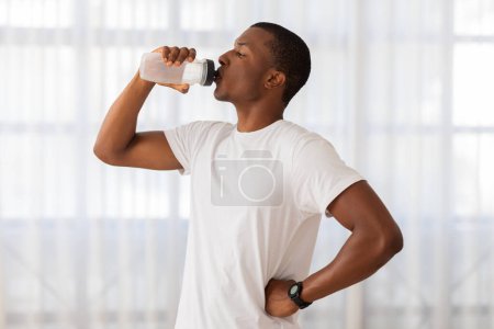 Téléchargez les photos : Un Afro-Américain en forme dans un t-shirt blanc et un pantalon sombre prend une pause pour s'hydrater avec une bouteille d'eau après l'exercice - en image libre de droit