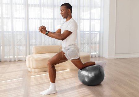 A determined black man concentrates while kneeling on a balance ball in a sunny, modern-looking room