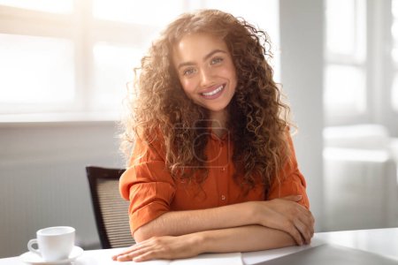 Téléchargez les photos : Jeune femme expose une pose détendue et réfléchie, le menton sur la main à son bureau avec un ordinateur portable et un café - en image libre de droit