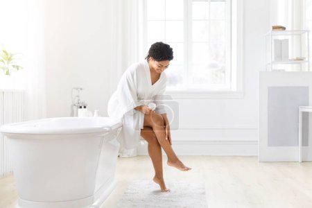 Mujer afroamericana en un albornoz se muestra comprobando su piel en la pierna, destacando una rutina de autocuidado