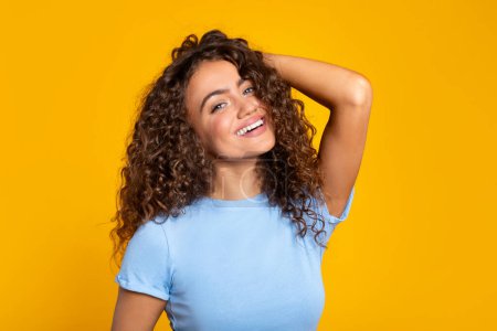 Cheerful curly-haired young woman in blue t-shirt smiling and touching her hair on a sunny yellow background