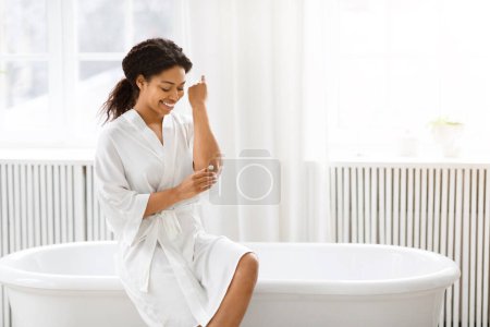 Téléchargez les photos : Contenu Femme afro-américaine dans un peignoir blanc se trouve sur le bord d'une baignoire dans une salle de bain propre et lumineuse, en utilisant la crème pour le corps - en image libre de droit