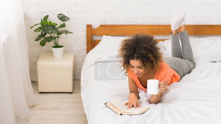 Photo for African-american girl reading book and drinking coffee in cozy bedroom, empty space - Royalty Free Image